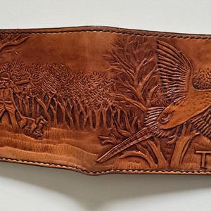 May include: A brown leather wallet with an embossed design of a pheasant in flight and a hunter in a forest. The wallet has the initials "TW" embossed on the bottom right corner.