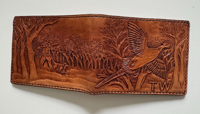 May include: A brown leather wallet with an embossed design of a pheasant in flight and a hunter in a forest. The wallet has the initials "TW" embossed on the bottom right corner.