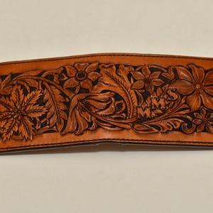 May include: A brown leather wallet with a floral design carved into the front. The design includes leaves, flowers, and the initials "M.M.S."