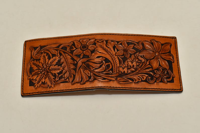 May include: A brown leather wallet with a floral design carved into the front. The design includes leaves, flowers, and the initials "M.M.S."