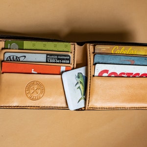 May include: A brown leather wallet with multiple card slots and a small pocket for cash. The wallet is open and contains several cards, including a Cabela's Conservation Card, a Costco membership card, and a roadside assistance card. The wallet also has a small, white card with a picture of a fish on it.