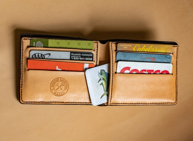 May include: A brown leather wallet with multiple card slots and a small pocket for cash. The wallet is open and contains several cards, including a Cabela's Conservation Card, a Costco membership card, and a roadside assistance card. The wallet also has a small, white card with a picture of a fish on it.