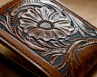 May include: A brown leather wallet with a floral design embossed on the front. The design is intricate and detailed, with a large flower in the center and smaller leaves and vines surrounding it.