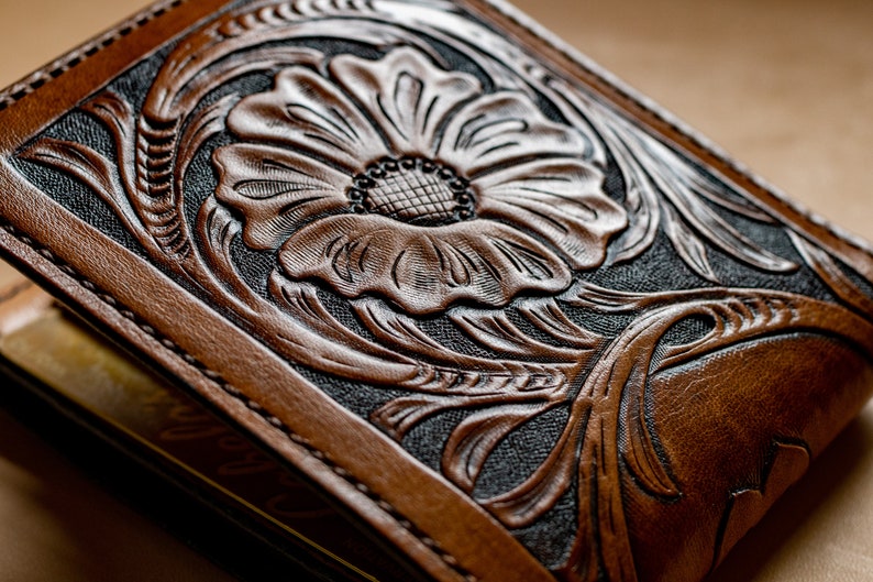 May include: A brown leather wallet with a floral design embossed on the front. The design is intricate and detailed, with a large flower in the center and smaller leaves and vines surrounding it.