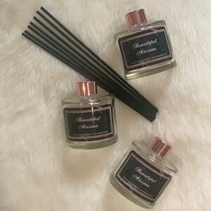 May include: Three glass bottles with black labels that read "Beautiful Aroma" and rose gold caps. The bottles are filled with a clear liquid and are sitting on a white fluffy surface. Black diffuser sticks are in the background.