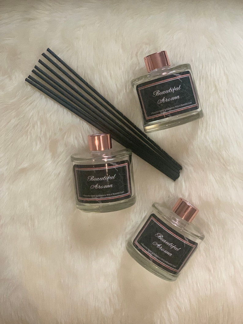 May include: Three glass bottles with black labels that read "Beautiful Aroma" and rose gold caps. The bottles are filled with a clear liquid and are sitting on a white fluffy surface. Black diffuser sticks are in the background.