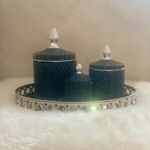 May include: A set of three black, textured glass jars with silver lids, presented on a silver tray with crystal detailing. The jars vary in size and are placed on a white, fluffy surface.