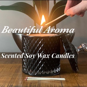 Puede incluir: Un tarro de vela de vidrio negro con una tapa plateada y un diseño decorativo. La vela está encendida y el tarro está sobre una superficie de madera. El texto "Beautiful Aroma Scented Soy Wax Candles" es visible en la imagen.