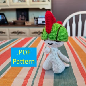 May include: A handmade crochet doll with a white body, green head, and red accents. The doll is standing on a striped tablecloth. The image includes the text ".PDF Pattern" in a blue box.