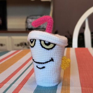 May include: A white crocheted cup with a pink straw and a yellow star. The cup has a cartoon face with eyes, a smile, and a straw sticking out of the top.