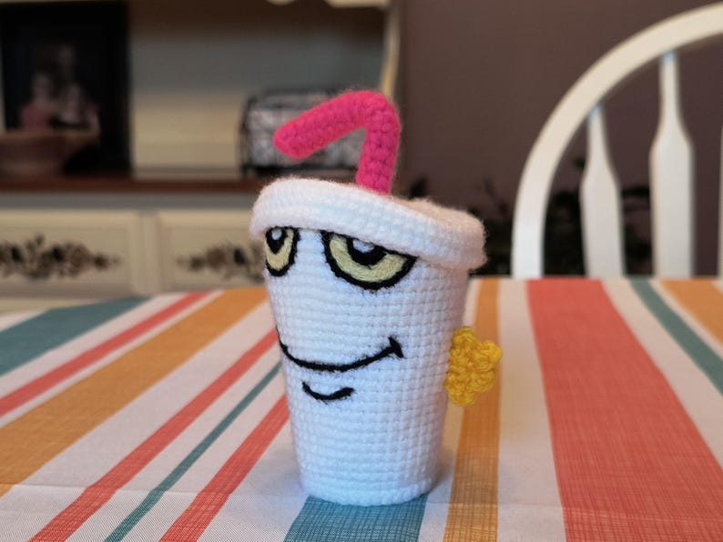 May include: A white crocheted cup with a pink straw and a yellow star. The cup has a cartoon face with eyes, a smile, and a straw sticking out of the top.