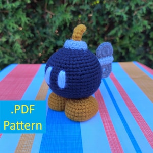 Puede incluir: Personaje de bomba de ganchillo hecho a mano en azul marino, blanco y dorado. El personaje tiene ojos ovalados blancos, una mecha amarilla y pies dorados. Un ala gris pequeña está unida al costado. El texto ".PDF Pattern" está en un cuadro azul.