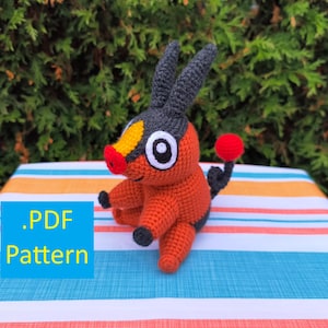 May include: A crocheted orange and black creature with large white eyes, a red nose, and a red ball tail. The creature has black ears and is sitting on a striped surface. The text ".PDF Pattern" is in a blue box.