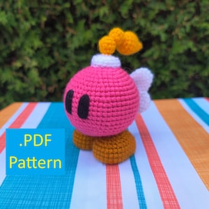 Puede incluir: Un personaje de bomba de ganchillo hecho a mano en rosa, con ojos negros, alas blancas y llamas amarillas. El personaje está sentado sobre una superficie a rayas. La imagen incluye el texto ".PDF Pattern" en un cuadro azul.