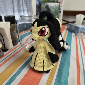 May include: A crocheted yellow and black Pokemon plush toy with red eyes. The toy is sitting on a striped table cloth.