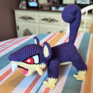 May include: A purple and yellow crocheted plush toy resembling a cartoon animal with a curled tail. The toy has a fierce expression with red eyes and sharp teeth.