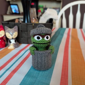 Puede incluir: Un títere de crochet verde y gris de un personaje que sostiene un cubo de basura de crochet gris. El títere tiene grandes ojos verdes y un sombrero gris. El títere está sentado en un mantel a rayas.