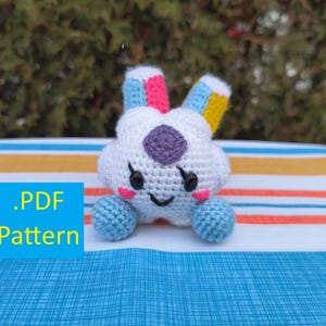 Könnte beinhalten: Ein weißes, gehäkeltes, wolkenförmiges Spielzeug mit blauen, rosa und gelben Akzenten. Es hat eine lila Mitte, rosa Wangen und ein lächelndes Gesicht. Der Text ".PDF Pattern" befindet sich in einem blauen Quadrat unten links. Das Spielzeug liegt auf einer gestreiften Oberfläche.