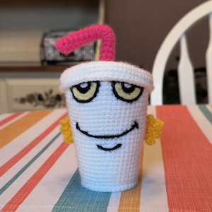 Puede incluir: Una taza blanca de ganchillo con una pajita rosa y ojos amarillos. La taza tiene una cara sonriente y está sentada sobre un mantel a rayas.