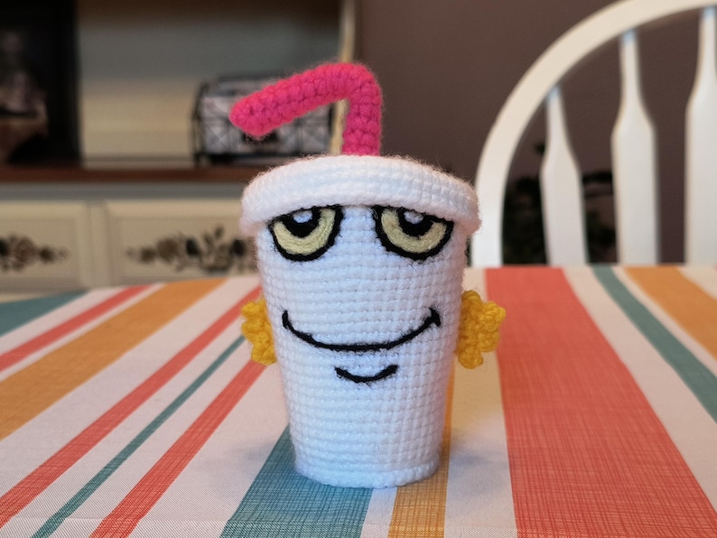 May include: A white crocheted cup with a pink straw and yellow eyes. The cup has a smiling face and is sitting on a striped tablecloth.
