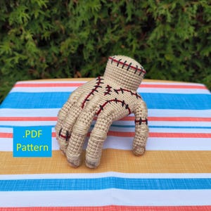 May include: A beige crochet hand with red and black stitching, resting on a striped surface. The hand is detailed with stitches and has a textured appearance. The text ".PDF Pattern" is in a blue box.