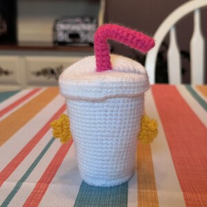 May include: A crocheted white cup with a pink straw and yellow accents. The cup is sitting on a striped table cloth.