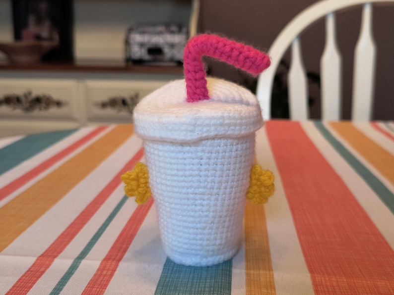 May include: A crocheted white cup with a pink straw and yellow accents. The cup is sitting on a striped table cloth.