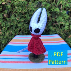 May include: A handmade crochet figure of a character with a white head, black eyes, and a red cape. The figure is on a green base, with a gray sword. The text ".PDF Pattern" is in a blue box.