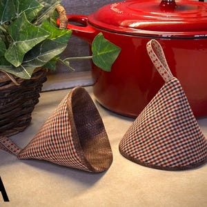 May include: Two cone-shaped fabric pot holders featuring a small red, black, and cream checkered pattern with a brown interior lining and a small patterned hanging loop on each. The letter A is printed in the bottom left corner of the image.