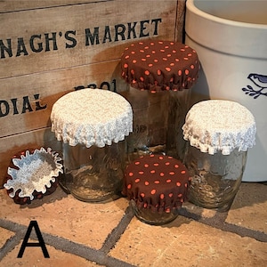 May include: A collection of clear glass jars with fabric covers featuring elasticized ruffled edges. The covers come in two patterns: a brown fabric with small orange polka dots and a light beige fabric with a fine, intricate floral print.