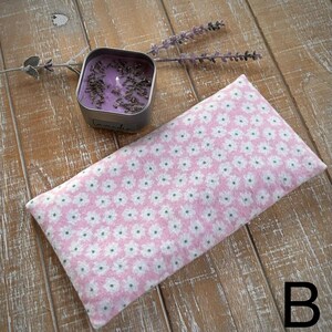 Op de afbeelding: Een roze oogkussen met bloemenpatroon en een kleine vierkante blikje met lavendelgeurkaars liggen op een houten oppervlak. Het oogkussen heeft een herhalend patroon van witte bloemen. Een takje lavendel ligt in de buurt van de kaars.