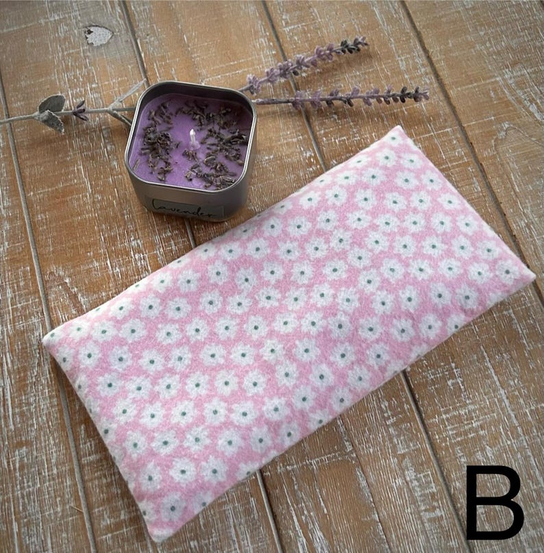 Op de afbeelding: Een roze oogkussen met bloemenpatroon en een kleine vierkante blikje met lavendelgeurkaars liggen op een houten oppervlak. Het oogkussen heeft een herhalend patroon van witte bloemen. Een takje lavendel ligt in de buurt van de kaars.
