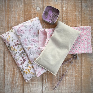 Op de afbeelding: Een assortiment lavendel-thema items op een houten oppervlak. Inclusief een rechthoekig beige stoffen kussen, verschillende bloemenstoffen zakjes in roze en wit, en een kleine metalen blik met een paarse kaars en gedroogde lavendel. Een takje lavendel is ook aanwezig.