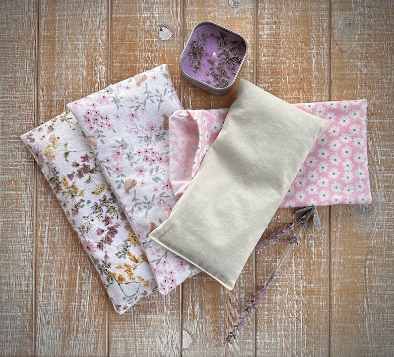 Op de afbeelding: Een assortiment lavendel-thema items op een houten oppervlak. Inclusief een rechthoekig beige stoffen kussen, verschillende bloemenstoffen zakjes in roze en wit, en een kleine metalen blik met een paarse kaars en gedroogde lavendel. Een takje lavendel is ook aanwezig.