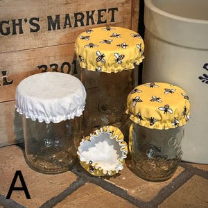 May include: Four glass mason jars with decorative fabric jar covers. Three jars have yellow covers with black and white bee patterns. One jar has a white cover with small white polka dots. The jars are on a brick surface.