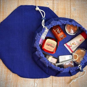 May include: A deep blue drawstring makeup bag, open to reveal various cosmetic products. The bag is made of a textured fabric and has a matching lid. Inside are compacts, tubes, and bottles of makeup. The bag is on a light wood surface.
