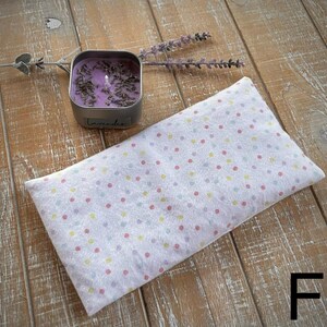 Op de afbeelding: Een wit, rechthoekig oogkussen met een pastelkleurig stippenpatroon ligt op een houten oppervlak. Een kleine, vierkante metalen blikken kaars met het woord "lavendel" staat naast het kussen. Er zijn ook gedroogde lavendel takjes aanwezig.