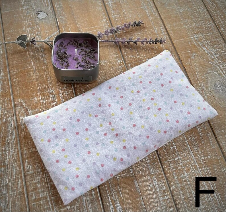 Op de afbeelding: Een wit, rechthoekig oogkussen met een pastelkleurig stippenpatroon ligt op een houten oppervlak. Een kleine, vierkante metalen blikken kaars met het woord "lavendel" staat naast het kussen. Er zijn ook gedroogde lavendel takjes aanwezig.