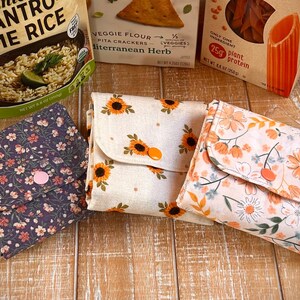 May include: Three square fabric pouches with fold-over flaps secured by plastic snaps. The left pouch is dark purple with small pink and white flowers. The center pouch is cream with sunflowers. The right pouch is white with orange and green floral prints.