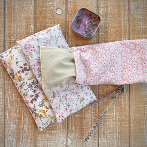Op de afbeelding: Een verzameling bloemmotief oogkussens in roze, wit en beige, samen met een kleine tinnen kaars gevuld met lavendel en een takje lavendel. De kussens hebben verschillende bloemmotieven, wat een rustgevende uitstraling cre&euml;ert.