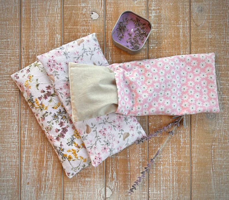 Op de afbeelding: Een verzameling bloemmotief oogkussens in roze, wit en beige, samen met een kleine tinnen kaars gevuld met lavendel en een takje lavendel. De kussens hebben verschillende bloemmotieven, wat een rustgevende uitstraling cre&euml;ert.