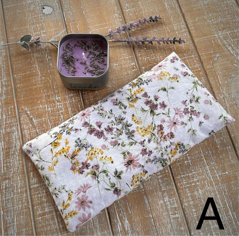 Op de afbeelding: Een rechthoekig kussen met een bloemenpatroon in wit, paars, geel en groen. Daarnaast een kleine, vierkante zilveren kaarsenpot met een paarse kaars en lavendelknopjes. Op de pot staat "Lavender".