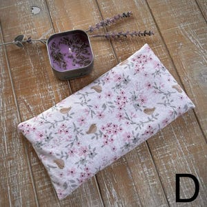 Op de afbeelding: Een rechthoekig oogkussen met een bloemenpatroon van roze bloemen en kleine vogelmotieven. Een kleine, vierkante metalen kaars met lavendel staat ernaast. De achtergrond is een houten oppervlak.