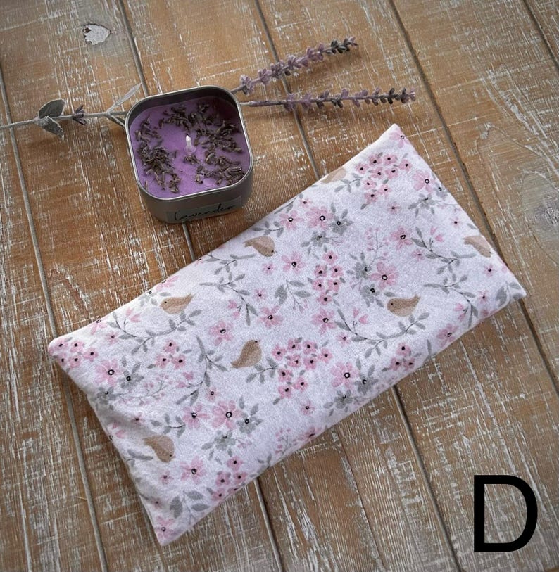Op de afbeelding: Een rechthoekig oogkussen met een bloemenpatroon van roze bloemen en kleine vogelmotieven. Een kleine, vierkante metalen kaars met lavendel staat ernaast. De achtergrond is een houten oppervlak.