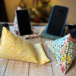 May include: Several fabric phone stands in various floral patterns and colors are displayed on a wooden surface. The stands are triangular, designed to prop up a phone. One stand is yellow with small floral details.
