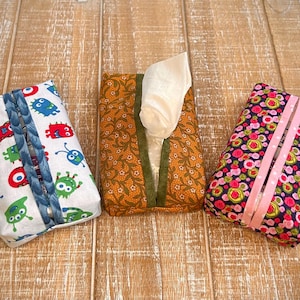 May include: Three fabric tissue box covers. One has a white background with colorful cartoon monsters, one has a brown background with a floral pattern, and one has a dark blue background with pink and green circles.