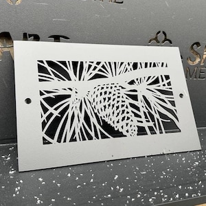 Puede incluir: Una rejilla de ventilación de metal gris con un diseño recortado de una piña y agujas de pino. La rejilla de ventilación es rectangular y tiene esquinas redondeadas. El diseño es blanco y el fondo es gris.