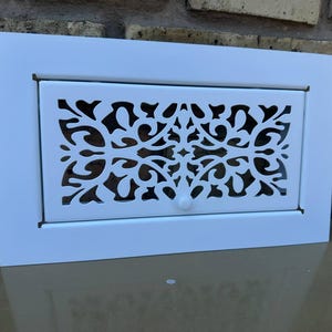 Könnte beinhalten: Ein weißer, rechteckiger Lüftungsdeckel mit einem dekorativen, durchbrochenen Design. Die Mittelplatte weist ein kompliziertes Blumenmuster auf, und ein kleiner Knopf ist unten sichtbar. Der Lüftungsdeckel ist vor einer Backsteinmauer angebracht.