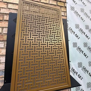 Pode incluir: Tela retangular de metal dourado com um padrão geométrico. A tela é exibida contra uma parede de tijolos e uma faixa que diz "SAVA ART METAL". A tela tem um design decorativo.