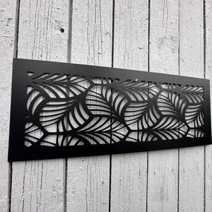 May include: A black metal decorative panel with a leaf design. The panel is rectangular and features a cut-out pattern of stylized leaves. It is mounted on a weathered, white-painted wooden surface.